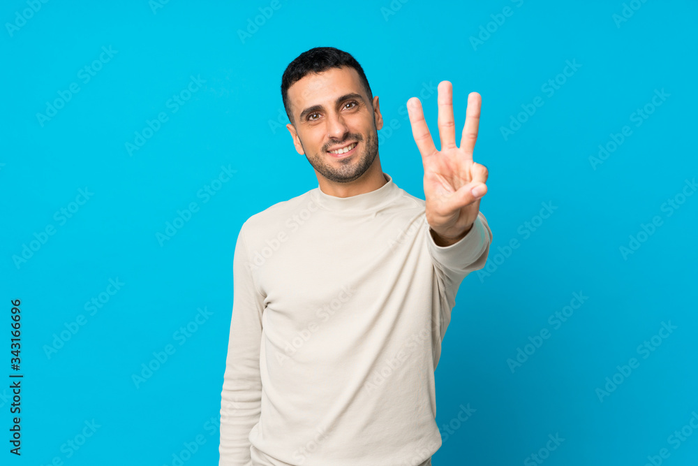 Young man over isolated blue background happy and counting three with fingers Young man over isolated blue background happy and counting three with fingers
