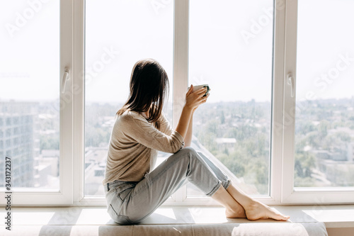 Young woman sitting on a windowsill by the window with a city view, drinking coffee or tea in the morning.