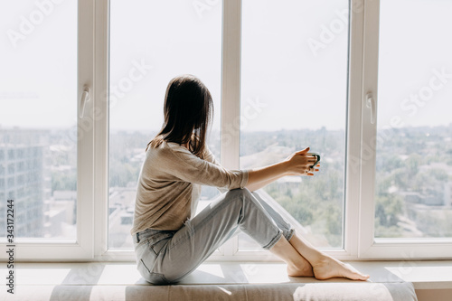 Young woman looking through the window with a city view, sitting on a windowsill, drinking coffee or tea in the morning.