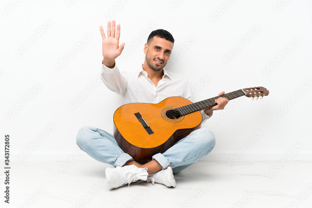Obraz premium Young man sitting on the floor with guitar saluting with hand with happy expression