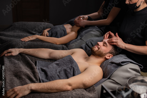 happy married couple lying on massage desks in spa salon, man and woman get cosmetic beauty procedure, take care of skin on head and face, face ligt anti aging massage by professionals