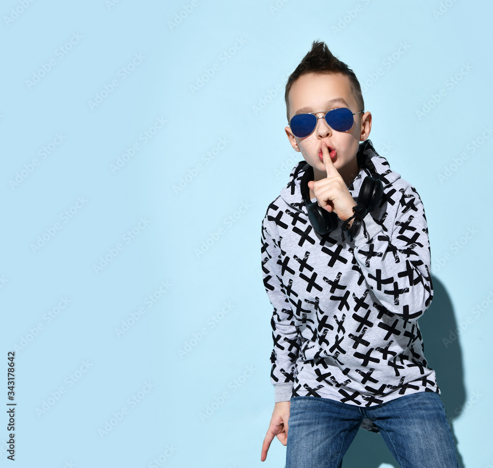 Fototapeta premium Teenage child with headphones on neck, in sunglasses, hoodie, black bracelet. He showing be quiet sign, posing on blue background