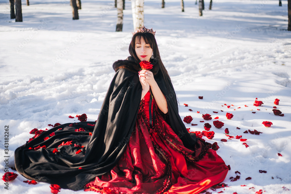 Naklejka premium A beautiful girl with long black hair is sitting on the white snow in a red dress and cloak. Princess sits with a rose in a crown with precious stones. Fantasy photography