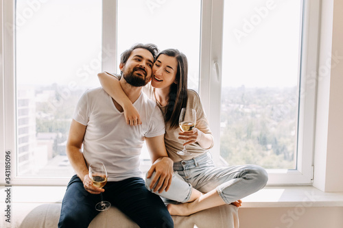 Young couple of man and woman in casual clothes, sitting on windowsill in day light hugging and drinking wine.