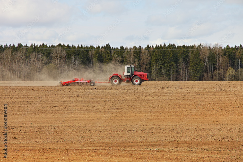 Obraz premium Red big wheeled tractor with large plough ploughs the land in a field on forest background at spring day, rural farm landscape