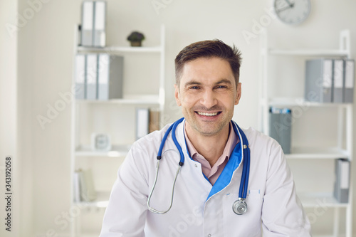 Male doctor with a stethoscope smiling while sitting at a table in a white interior of a clinic office.