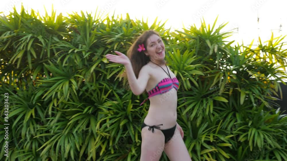 lady in bikini with purple flower in long loose flowing hair jumps and waves near tropical plants at back bright summer sunlight lady in bikini with purple flower in long loose flowing hair jumps and waves near tropical plants at back bright summer sunlight