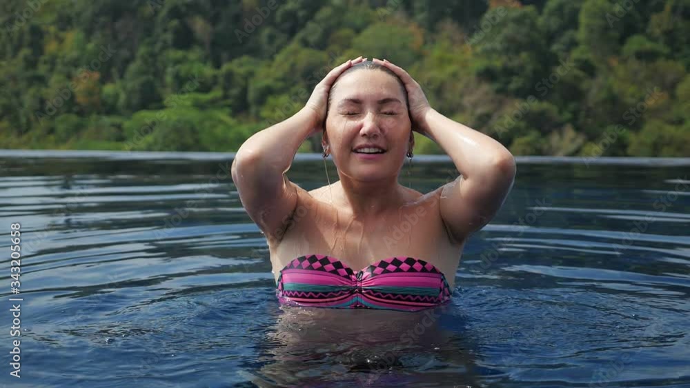 lady in bikini fixes black hair with hands after surfacing from hotel swimming pool water against green plants slow motion close view lady in bikini fixes black hair with hands after surfacing from hotel swimming pool water against green plants slow motion close view