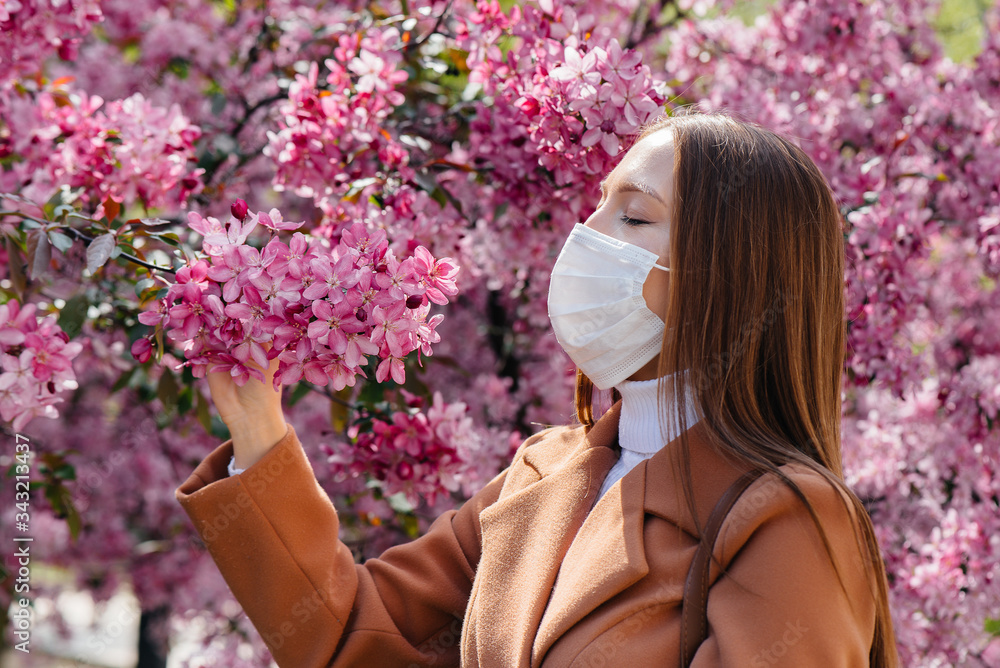 Fototapeta premium A young girl takes off her mask and breathes deeply after the end of the pandemic on a Sunny spring day, in front of blooming gardens. Protection and prevention covid 19