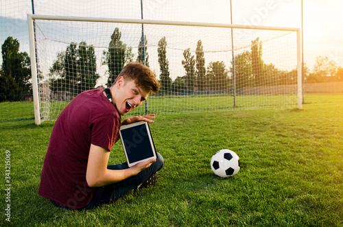 Ecstatic boy holding tablet while sitting on the grass of football stadium 
