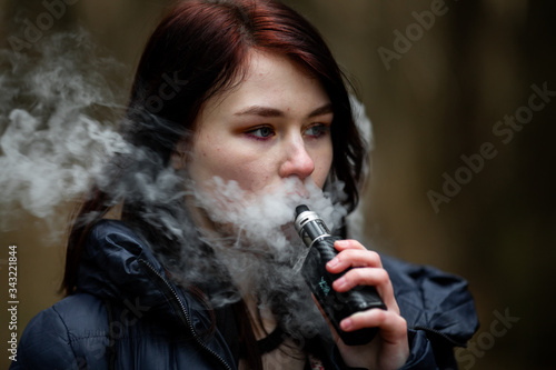 Vape teenager. Young pretty caucasian brunette girl smoking an electronic cigarette on the street in the spring. Deadly bad habit.