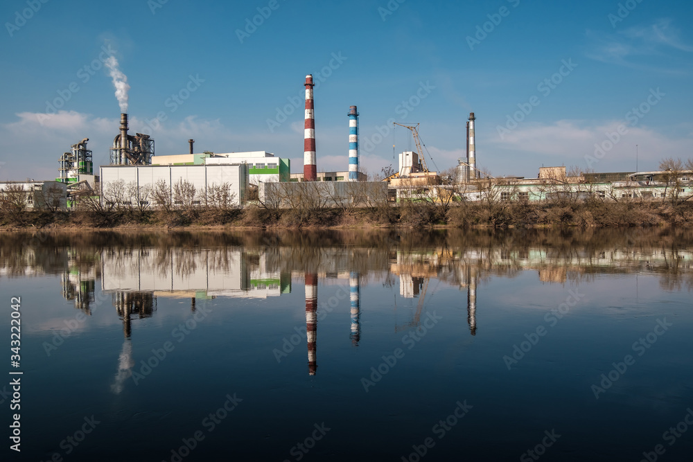 Obraz premium pipes of woodworking enterprise plant sawmill near river. Air pollution concept. Industrial landscape environmental pollution waste of thermal power plant