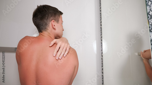 Tablou pe pânză handsome guy with bare torso looks at sunburned body in mirror standing in bathr