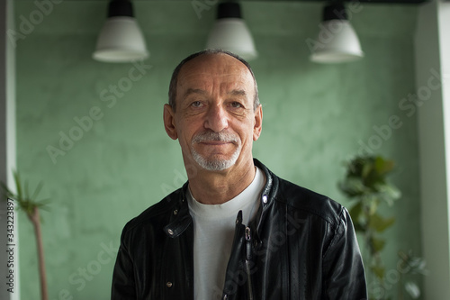 Portrait of senior mature man in leather jacket sitting at the sofa and looking at the camera on green studio background