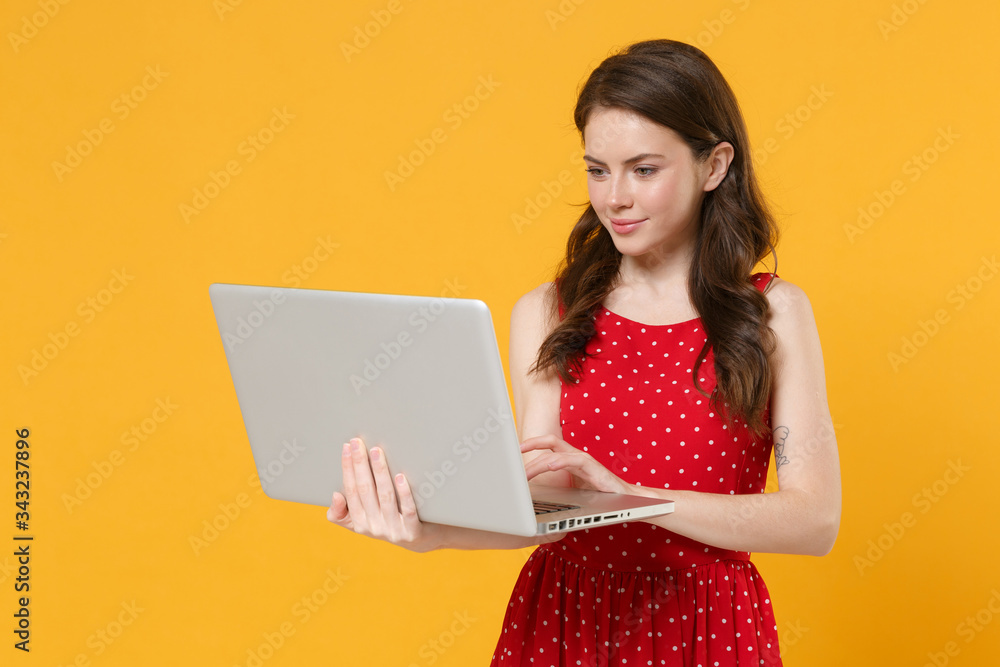 Naklejka premium Smiling young brunette woman girl in red summer dress posing isolated on yellow wall background studio portrait. People emotions lifestyle concept. Mock up copy space. Working on laptop pc computer.