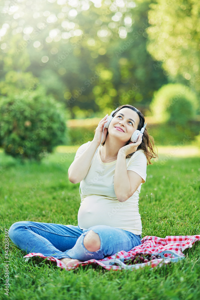Happy young pregnant woman sitting on carpet and listening music on headphones from smartphone at park. Pregancy, Relaxation and Health care concept. Happy young pregnant woman sitting on carpet and listening music on headphones from smartphone at park. Pregancy, Relaxation and Health care concept.