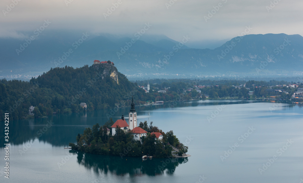 Church of pilgrimage Mariä Himmelfahrt on island in Lake Bled, during cloudy sunrise, castle in background, Bled Slovenia.