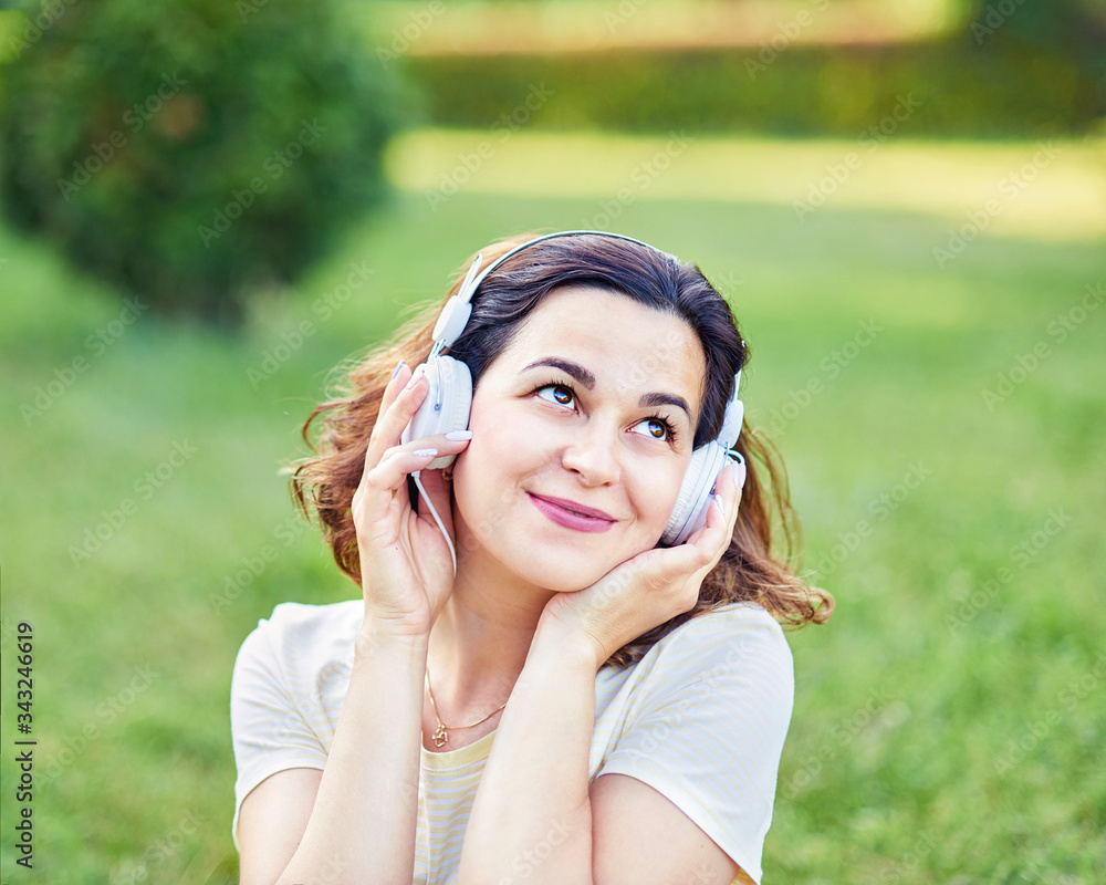 Happy young pregnant woman sitting on carpet and listening music on headphones from smartphone at park. Pregancy, Relaxation and Health care concept.
