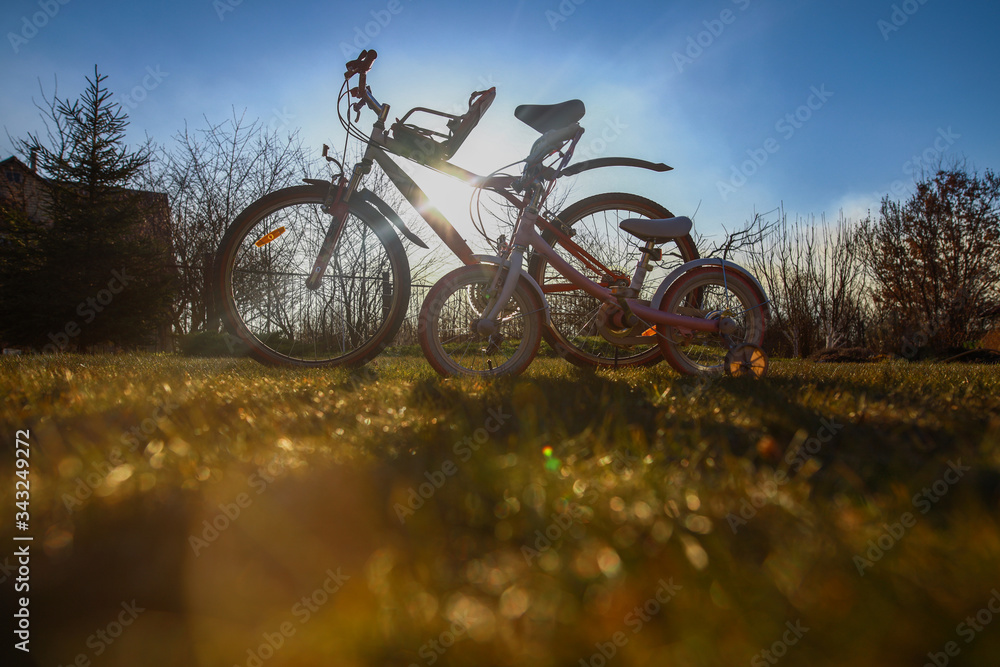 Fototapeta premium Two bicycles - adult and kid's one - standing outside in beautiful sunlight on green grass lane, bottom view, togetherness concept, kids bike seat