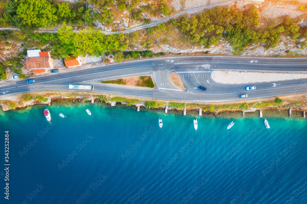 Aerial view of road in beautiful green forest and boats in the sea at sunset in summer. Colorful landscape with asphalt roadway, blue water, trees. Top view from drone of highway in Croatia. Travel