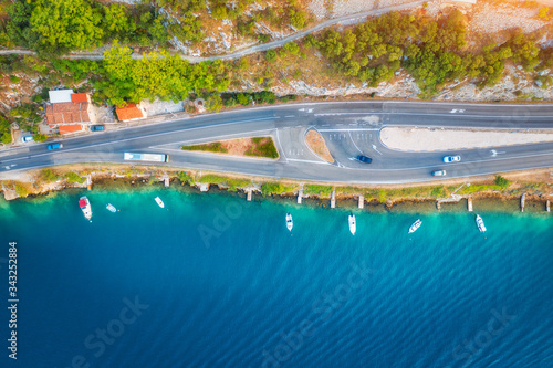 Aerial view of road in beautiful green forest and boats in the sea at sunset in summer. Colorful landscape with asphalt roadway, blue water, trees. Top view from drone of highway in Croatia. Travel