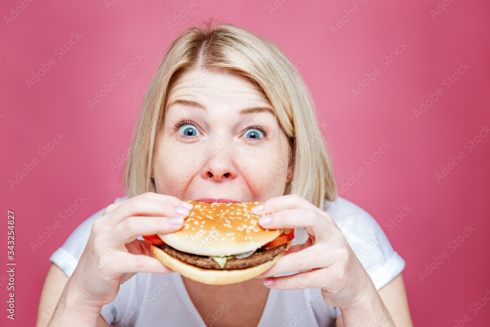 Blond woman eating a big juicy hamburger. Junk food and unhealthy lifestyle. Pink background. Close-up.