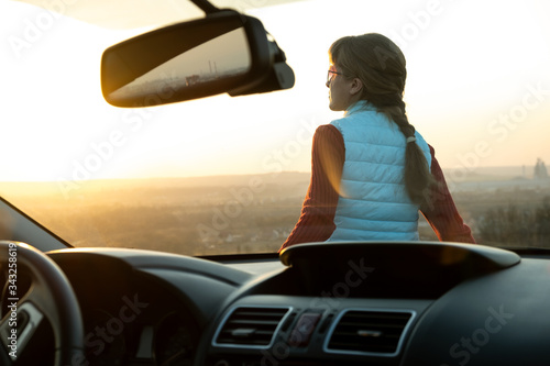 View from inside of a young woman standing near her car enjoying warm sunset view. Girl traveler leaning on vehicle hood looking at evening horizon. © bilanol