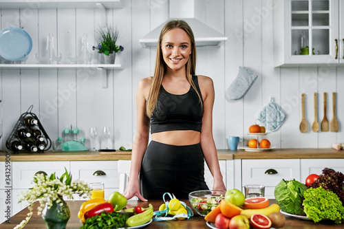 A sporty girl stands in the kitchen near a table with fruits and other useful foods. Healthy eating concept