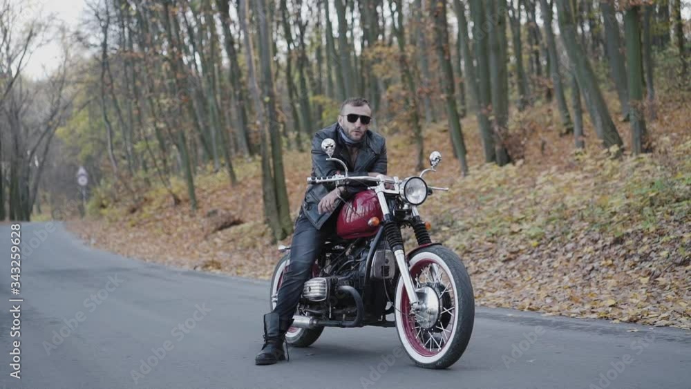 Portrait of brutal biker in sunglasses and leather on motorcycle on roadway Portrait of brutal biker in sunglasses and leather on motorcycle on roadway