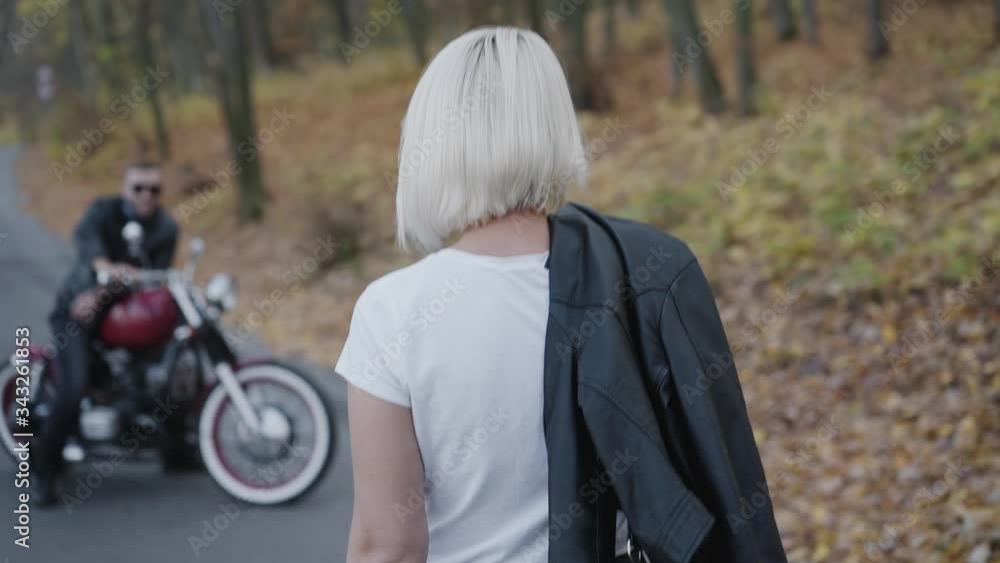 Back look of blonde comes to her smiling brutal biker with motorcycle in park