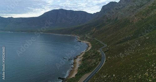 A striking aerial over cars speeding along a winding road at the feet of lush hills and rocky cliffs on the shore of a large bay - Cape Town, South Africa
