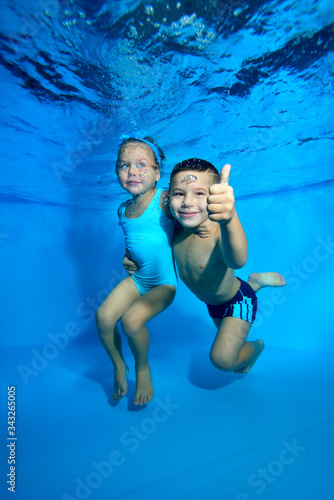 A little boy and his younger sister pose underwater in a children's pool. The boy smiles broadly, looks at the camera, and gives a thumbs-up. Closeup. Digital photo.