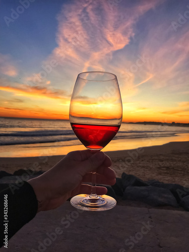 Woman man hand with a glass of red wine on a background of sunset sky and waves on the beach by the ocean in Portugal