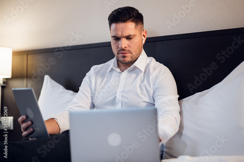 Modern job lifestyle. Serious concentrated man sitting on a bed at home, working on a laptop online, using internet