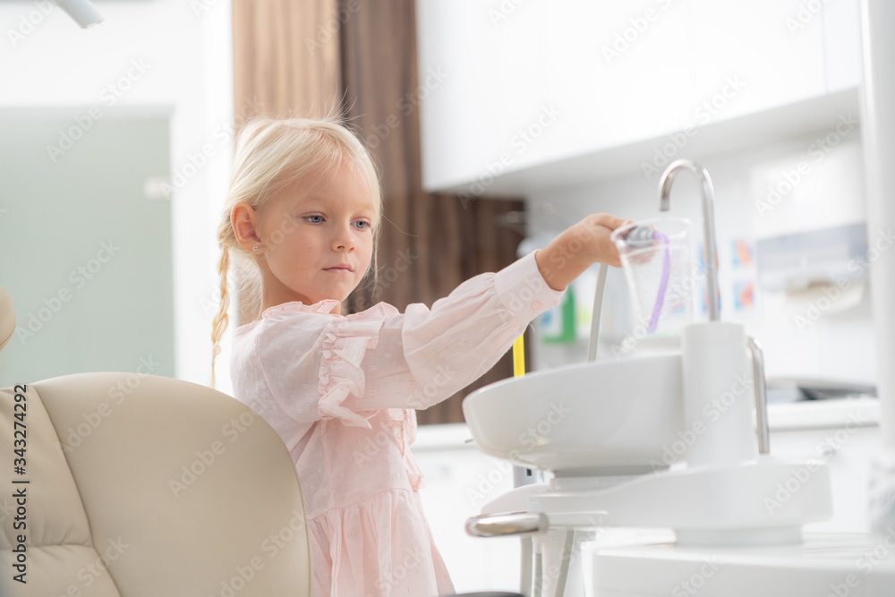 A little blond girl in the dental clinic. A cup with sterile water for rinsing the mouth in the foreground. A visit to a dentist concept A little blond girl in the dental clinic. A cup with sterile water for rinsing the mouth in the foreground. A visit to a dentist concept