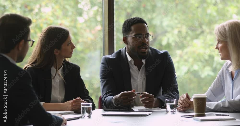 Serious african american businessman in eyeglasses holding negotiations with focused diverse partners in office. Motivated mixed race business people discussing project ideas at brainstorming meeting.