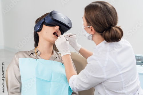 The dentist is doing her job, while the patient is lying with her mouth wide open, wearing VR glasses