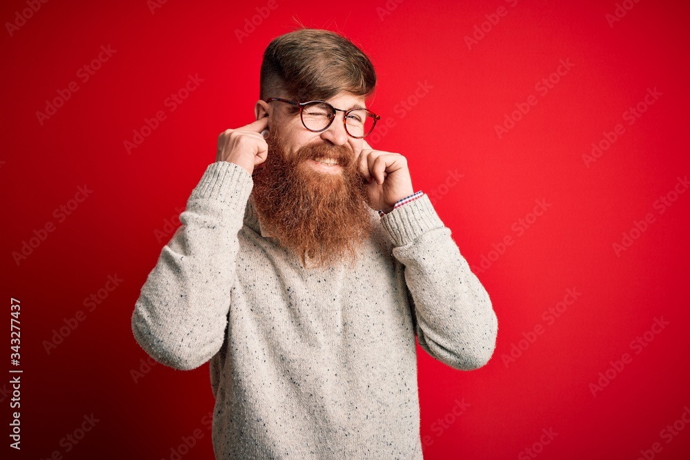 Handsome Irish redhead man with beard wearing casual sweater and glasses over red background covering ears with fingers with annoyed expression for the noise of loud music. Deaf concept. Handsome Irish redhead man with beard wearing casual sweater and glasses over red background covering ears with fingers with annoyed expression for the noise of loud music. Deaf concept.