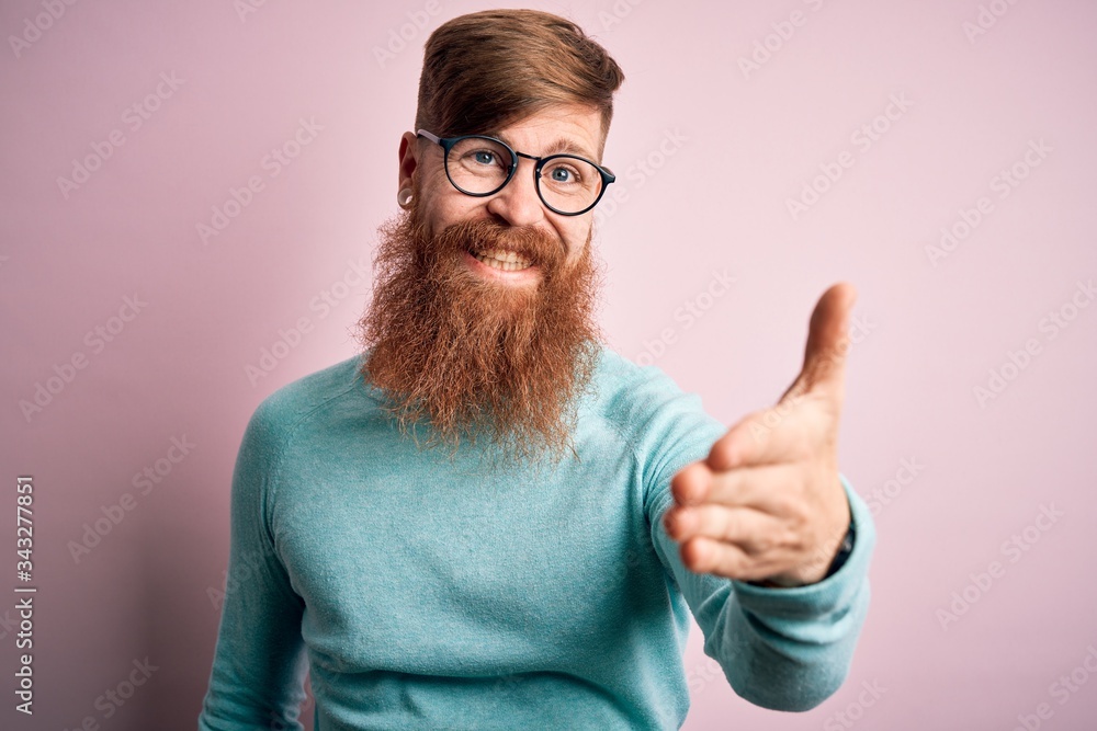 Handsome Irish redhead man with beard wearing glasses over pink isolated background smiling friendly offering handshake as greeting and welcoming. Successful business.