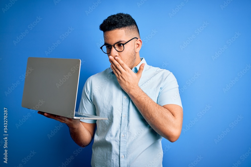 Young handsome businessman wearing glasses working using laptop over blue background cover mouth with hand shocked with shame for mistake, expression of fear, scared in silence, secret concept Young handsome businessman wearing glasses working using laptop over blue background cover mouth with hand shocked with shame for mistake, expression of fear, scared in silence, secret concept