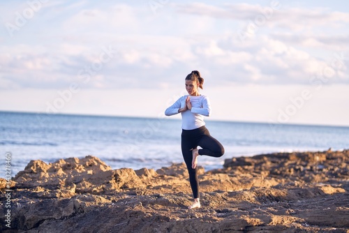 Fototapeta Naklejka Na Ścianę i Meble -  Young beautiful sportwoman smiling happy practicing yoga. Coach with smile on face teaching tree pose at the beach