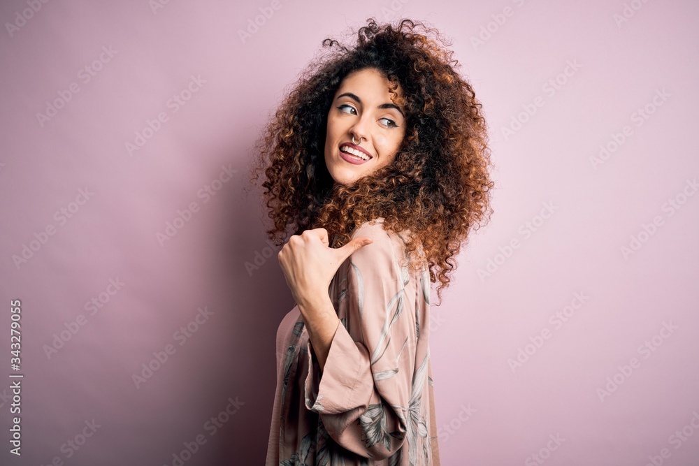 Young beautiful woman with curly hair and piercing wearing casual pink t-shirt smiling with happy face looking and pointing to the side with thumb up. Young beautiful woman with curly hair and piercing wearing casual pink t-shirt smiling with happy face looking and pointing to the side with thumb up.