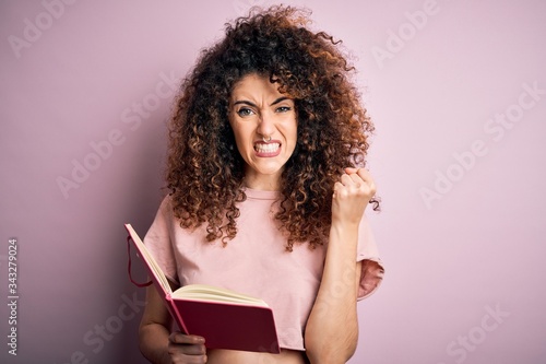 Young beautiful student woman with curly hair and piercing reading book over pink background annoyed and frustrated shouting with anger, crazy and yelling with raised hand, anger concept