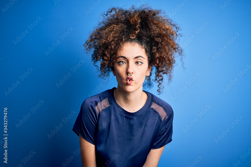 Young beautiful woman with curly hair and piercing wearing casual blue t-shirt making fish face with lips, crazy and comical gesture. Funny expression. Young beautiful woman with curly hair and piercing wearing casual blue t-shirt making fish face with lips, crazy and comical gesture. Funny expression.
