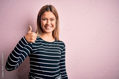 Young beautiful blonde woman wearing casual striped sweater over pink isolated background doing happy thumbs up gesture with hand. Approving expression looking at the camera showing success.