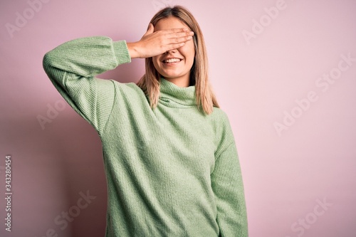 Young beautiful blonde woman wearing winter wool sweater over pink isolated background smiling and laughing with hand on face covering eyes for surprise. Blind concept. © Krakenimages.com