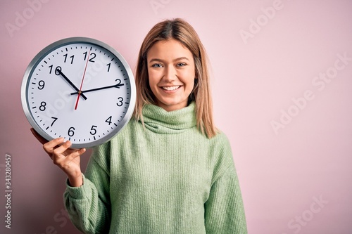 Young beautiful woman holding clock standing over isolated pink background with a happy face standing and smiling with a confident smile showing teeth