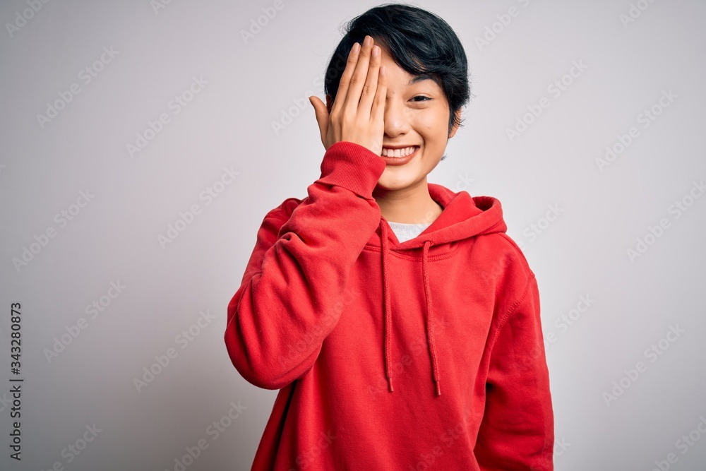 Young beautiful asian girl wearing casual sweatshirt with hoodie over white background covering one eye with hand, confident smile on face and surprise emotion. Young beautiful asian girl wearing casual sweatshirt with hoodie over white background covering one eye with hand, confident smile on face and surprise emotion.