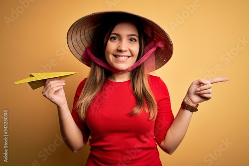 Tourist woman wearing traditional asian rice paddy straw hat holding paper plane for a trip very happy pointing with hand and finger to the side