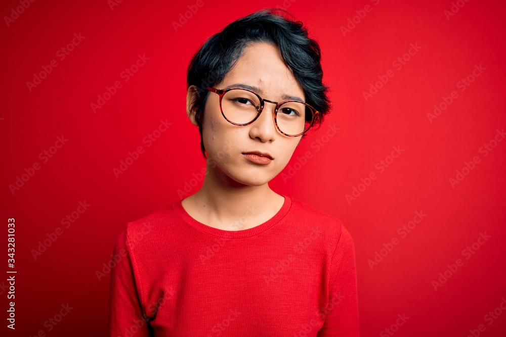 Young beautiful asian girl wearing casual t-shirt and glasses over isolated red background skeptic and nervous, frowning upset because of problem. Negative person. Young beautiful asian girl wearing casual t-shirt and glasses over isolated red background skeptic and nervous, frowning upset because of problem. Negative person.